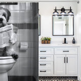 A black and white Cotton Cat shower curtain depicts a funny dog sitting on a toilet reading a newspaper. Adjacent is a modern white bathroom vanity with a dark faucet and light fixtures, creating the perfect setting for your quirky book reading dog décor.