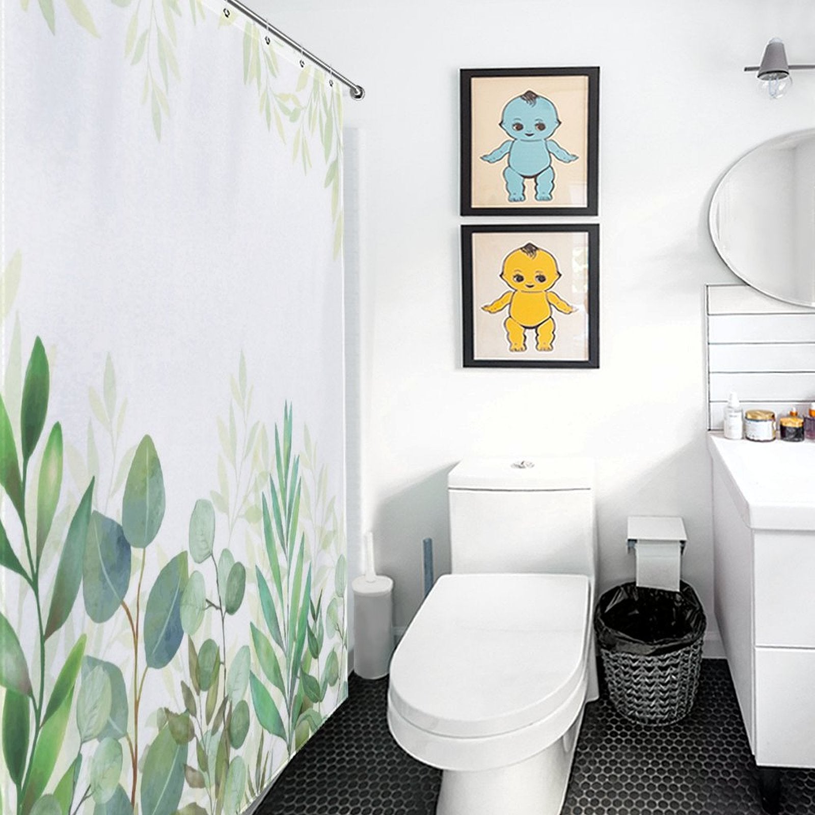 A modern bathroom with a white toilet, a circular mirror above a sink, and a Natural Modern Ombre Sage Green White Leaf Shower Curtain-Cottoncat by Cotton Cat. A waste bin sits nearby, and two framed artworks of colorful characters grace the wall, adding vibrant accents to the natural modern ombre design.