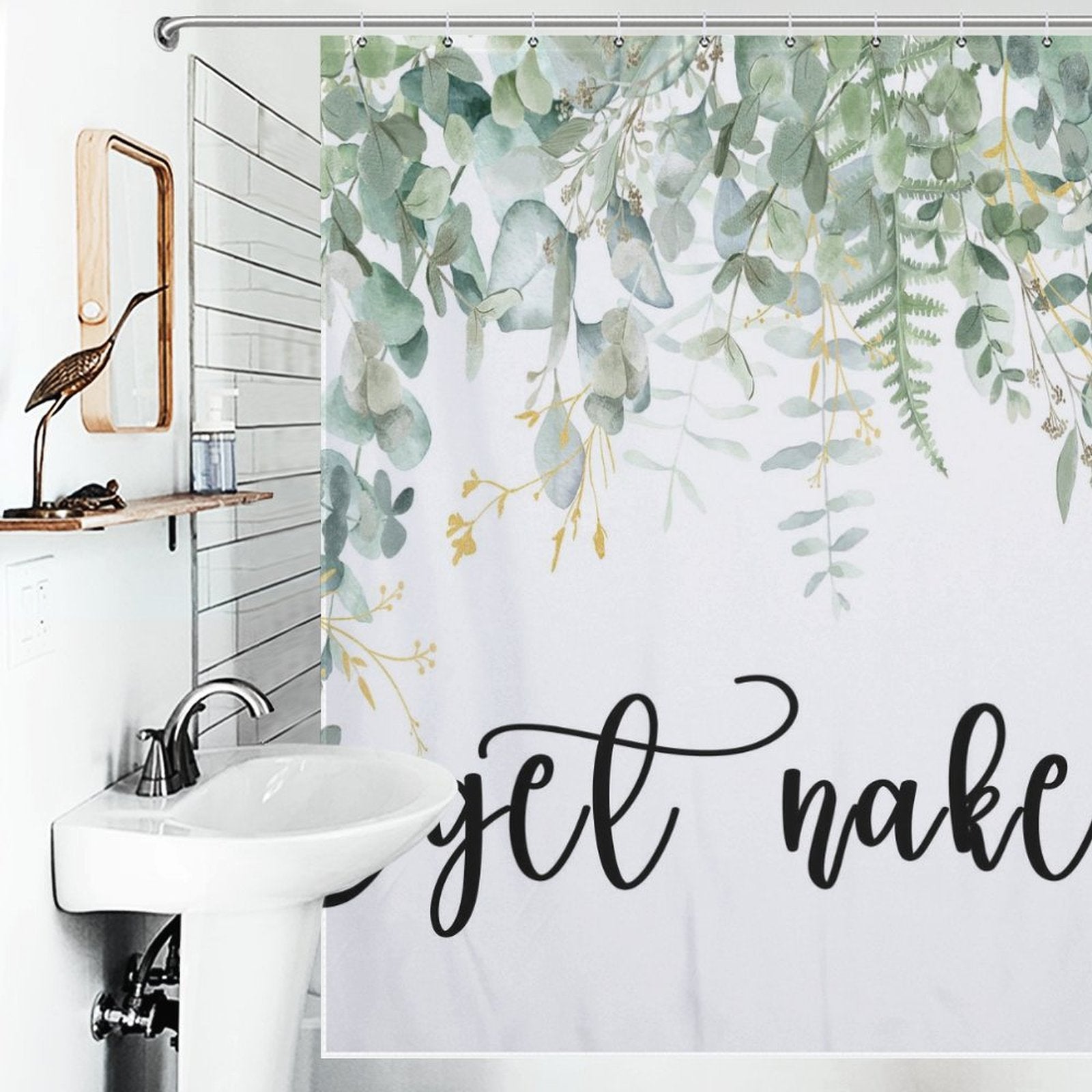 Bathroom with a white sink and a nature-themed shower curtain featuring eucalyptus leaves prints and the phrase "Get Naked" in cursive writing. A wooden bird sculpture is placed on a shelf beside the mirror.