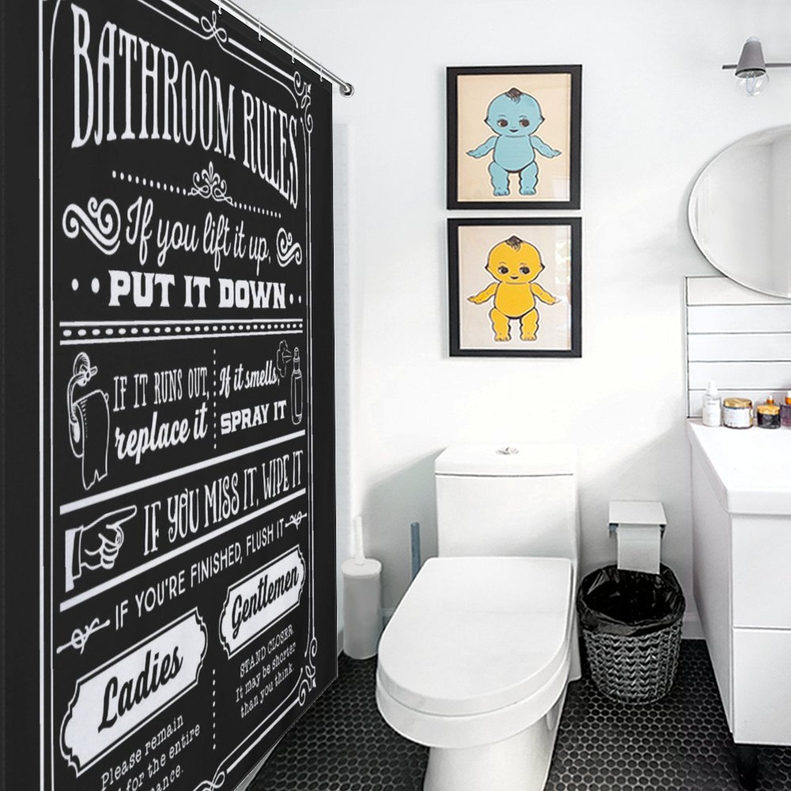 A bathroom with a white toilet, a sink, two framed cartoon character pictures on the wall, a trash bin, and a mirror above the sink features the Funny Quotes Shower Curtain Back and White Fable Motto -Cottoncat by Cotton Cat made from durable polyester fabric.