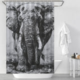 A Cotton Cat Black and White Elephant Shower Curtain elegantly hangs in a white bathroom. A white towel is draped over the bathtub, and toiletries are neatly arranged on a small table, completing the sophisticated bathroom decor.