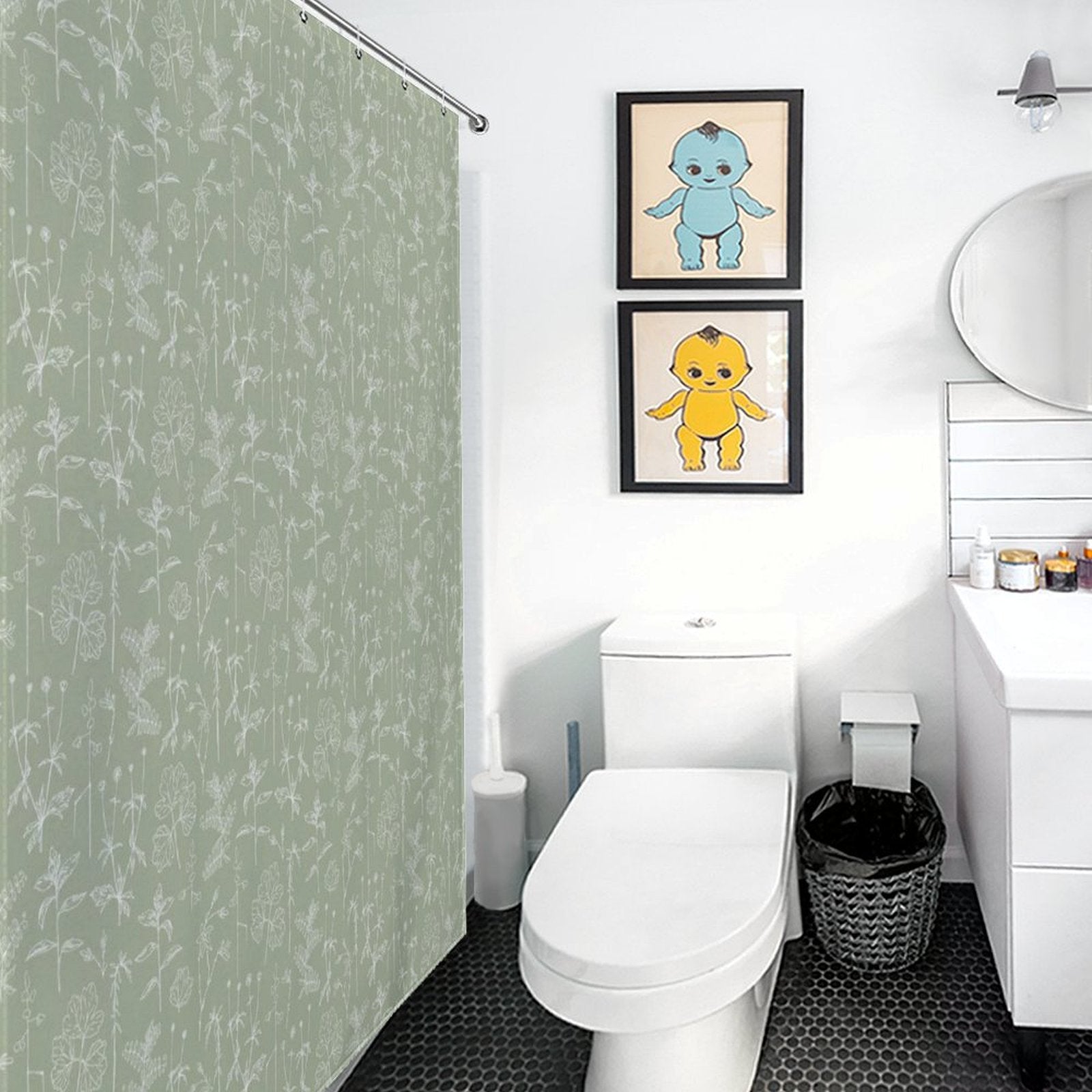 A clean bathroom with a Cotton Cat Boho Retro Sage Green Herbs Flower Shower Curtain showcasing a green floral design, white toilet, black trash can, and white sink. Two framed pictures of cartoon characters hang above the toilet. Round mirror above the sink.