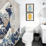A white bathroom features a toilet, a vanity with a sink, and a waste bin. The quirky bathroom decor includes the Funny Wave Monster Eating Cookies Shower Curtain-Cottoncat by Cotton Cat, and the wall is adorned with framed illustrations of blue and yellow characters.